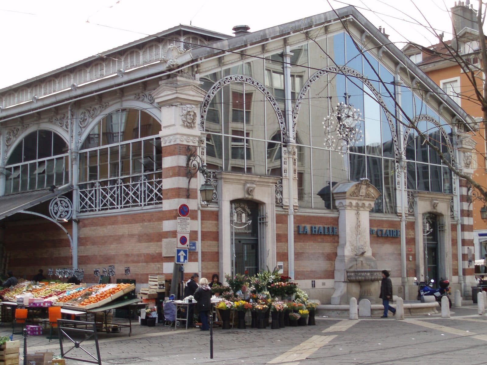 La halle Sainte-Claire vue de la place, avec les étals de fruits et de fleurs du marché de plein vent installés devant la façade de brique et de verre.